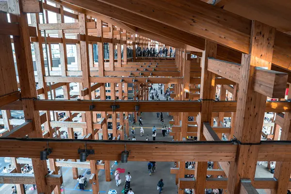 Kiriş gibi ızgaraları olan ahşap bir yapı, doğal ışığa izin veriyor. İnsanlar yürür ve aşağıda toplanır, halka açık bir etkinlik alanı olarak kullanıldığını vurgularlar.