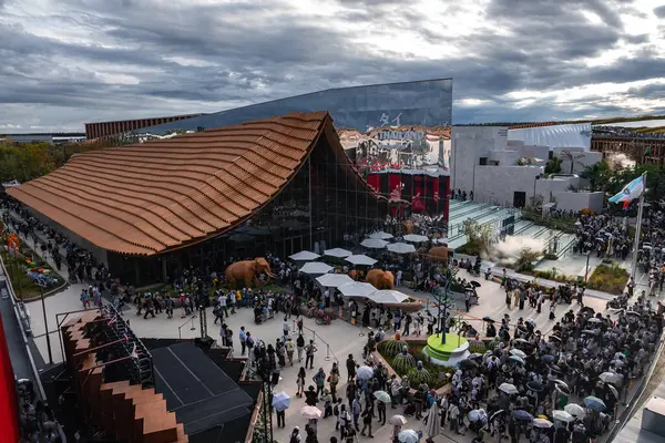Ziyaretçiler, Japonya 'nın Osaka kentindeki Dünya Fuarı 2025' te bulutlu bir gökyüzünün altında dalgalı bir terasotta çatısı ve cam duvarlı bir pavyonun yakınındaki sergileri keşfediyorlar..