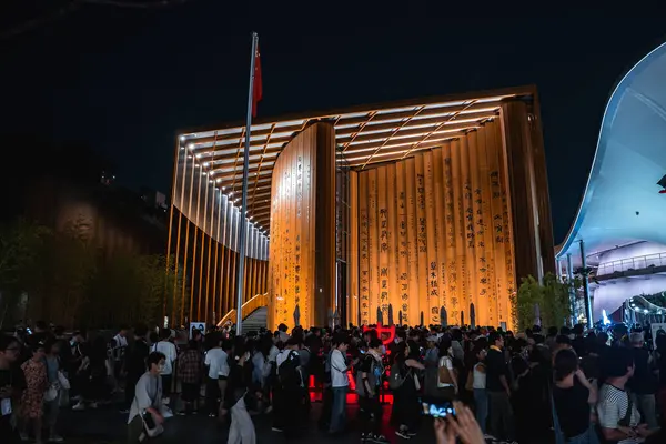 Dünya Fuarı 2025, Osaka 'da altın ışıklandırma ve hat panelleri olan eğri bir pavyon. Kırmızı bayrak ve canlı bir kalabalık coşkulu sahneyi canlandırıyor..