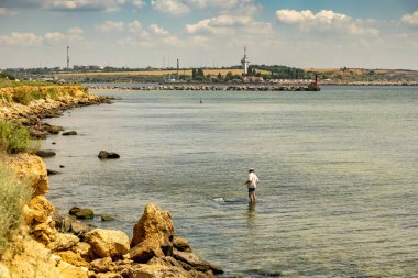 Yumuşak mavi gökyüzünün altında, sığ sulara odaklanmış bir balıkçı, kendi hattını çiziyor. Yumuşak dalgalar kayalık kıyıya çarpar ve sahilde huzurlu bir atmosfer yaratır..