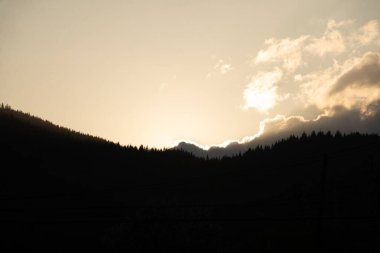 Gün geceye geçerken güneş ışığı bulutları delip geçer. Dağların zirvelerine sıcak bir parıltı yayar ve aşağıdaki yemyeşil ağaçları gölgeler..