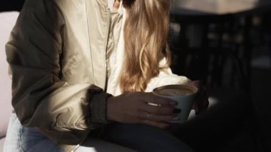 Güzel bir kız bankta otururken kahve içer. Kız dışarıda sıcak kahve içiyor. Güzel bir genç kadının portresi, yakın plan..