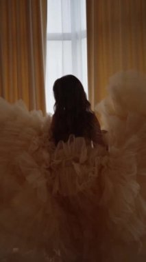 Uzun trenli, şenlikli elbiseli güzel bir kız pencerenin önünde duruyor..