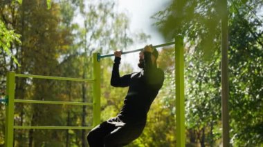 Spor kıyafetleri giymiş bir adam halka açık bir parkta yatay bir barda şınav çekiyor. Yürüyüş sırasında sporcunun etkinliği. Temiz havada fiziksel egzersizler.