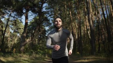 Genç sakallı bir atlet, bir çam ormanında ritmik olarak bir patika boyunca koşar. Spor kıyafetleriyle tatmin olmuş sporcu sabahları ormanda antrenman yapıyor.