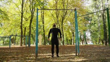 Bir adam kendini yatay bir çubuğa çekiyor. Açık hava sporları. Siyah pantolonlu ve tişörtlü bir sporcu spor sahasında barfiks çekiyor.