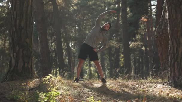 若い男が森の中で修行している 彼は体操をする 運動選手は訓練前に暖まる 健康的なライフスタイル — ストック動画