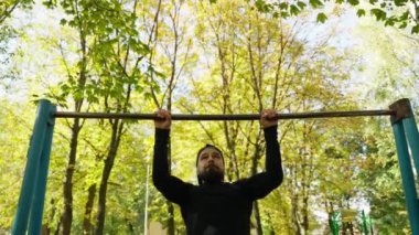 Atletik bir adam açık hava spor sahasında barfiks çekerek sırt kaslarını şişiriyor. Kaslı yakışıklı adam yatay çubukta antrenman yapıyor. Açık hava fitness ve vücut geliştirme konsepti.