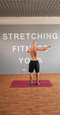 Handsome young man with bare torso and black shorts is doing sports at home on the internet. Active fitness man shows exercise for back and arm muscles with rubber band