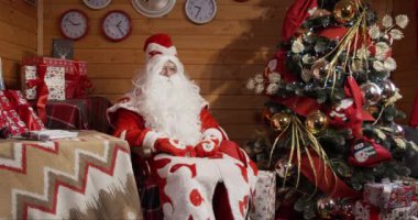 Santa Claus in a red suit, hat and gray beard is sitting in a chair in a decorated room near a glowing Christmas tree and giving presents.