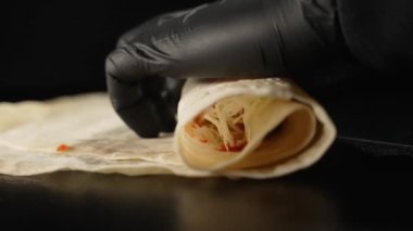 Male hands in black gloves of a restaurant chef wrap ingredients in pita bread in the process of preparing doner kebab. The cook prepares shawarma with meat and vegetables