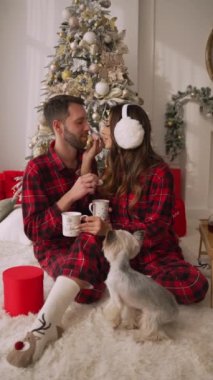 Happy couple drinking coffee together sitting on the floor at home. Sexy man and woman with teacups feeding each other sweet marshmallows