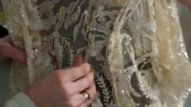 A woman fashion designer who creates a wedding dress to an exclusive order sews fabrics and rhinestones onto a dress worn by a mannequin. Jewelry work.