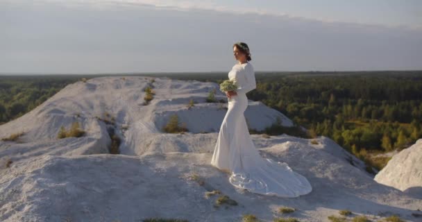 白いお祝いのドレスの花嫁は花の花束と山の上に立っている — ストック動画