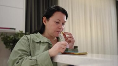 Happy woman reading message on cellphone and eating cookie while resting at home.