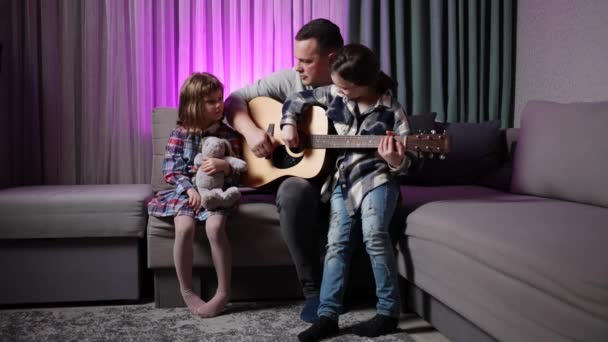 Padre Enseña Sus Hijas Tocar Guitarra Mientras Están Sentadas Sofá — Vídeo de stock
