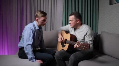 A man teaches his adult son to play the guitar while sitting on the couch at home