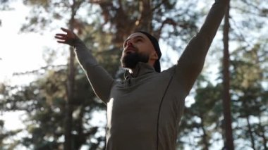 A young athletic guy is standing in the forest and doing breathing exercises. autumn leaves fall from the trees. Slow motion.