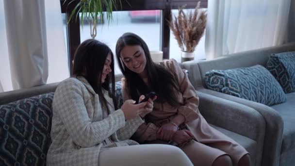 2人のファッショナブルな若い女性の友人がソファに座って リラックスし 笑顔で楽しく 携帯電話でオーディオメッセージを聞いて — ストック動画