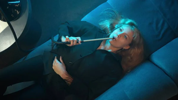 Seksi sarışın kız gece kulübünde kanepede uzanıyor ve nargile piposu içiyor. Genç bir kız nargile içmekten hoşlanır..