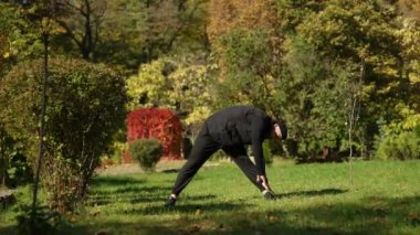 Genç bir adam bir sonbahar şehir parkında öne eğiliyor. Şehir parkında güneşli bir sonbahar günü. Sportif adam dışarıda yoga yapıyor..