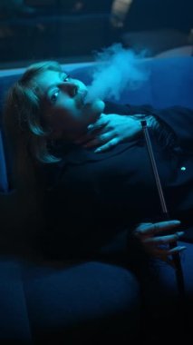 Genç seksi sarışın kızın yakın plan portresi nargile tüttürüyor ve kulüp kıyafetleriyle ağzından duman üflüyor..