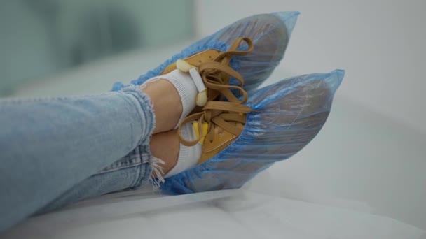 青いポリエチレンの靴カバーを持つ靴の女性の足 女性は足を組んで医療用のソファに横になっている — ストック動画