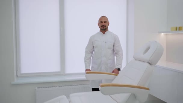 一个穿着白衣的年轻貌美的男人双臂交叉地站在办公室的内部 一位忙碌的全科医生在体检沙发旁边的医生办公室里摆姿势 — 图库视频影像