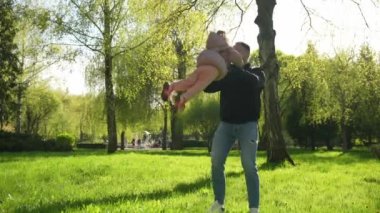 Yakışıklı genç bir adam küçük kızını kollarına alır, çocuk neşeyle güler. Yeşil bir parkta mutlu bir aile