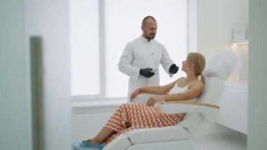 Bir kadın kozmetik kürsüsüne oturur, doktor gülümser, yanında durur ve sandalyeyi yattığı yere indirir.