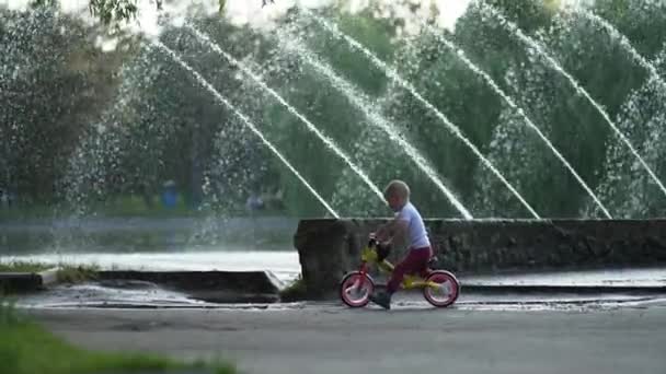 Niño Pequeño Monta Una Bicicleta Sin Pedales Cerca Una Fuente — Vídeos de Stock