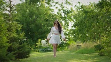 Büyük bir buket çiçekli mutlu bir esmer kız yeşil bir bahçede yürüyor.