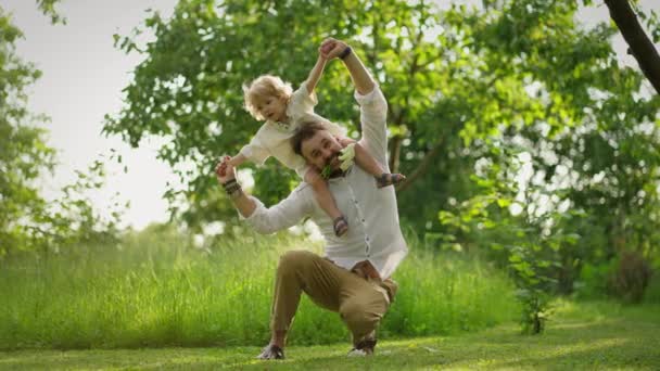 幸せな父親が肩に座っているかわいい小さな息子と一緒に歩いています 家族は草原で一緒に時間を過ごしています — ストック動画
