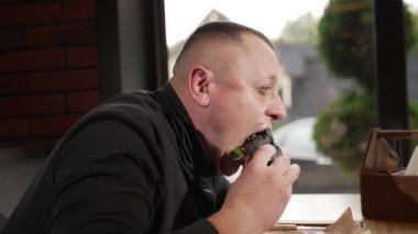 Kafede oturan bir adam sebze ve etle hamburger yiyor.