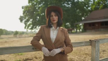 Güzel otlayan atların yanında duran makyajlı seksi kız. Ağır çekim. Gün batımında şapkalı seksi bir binicinin portresi.