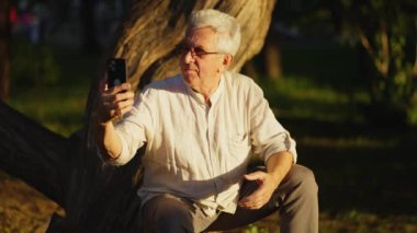 Yaşlı, kır saçlı bir adam güneş batarken bir ağacın yanında otururken cep telefonuyla fotoğrafını çekiyor.