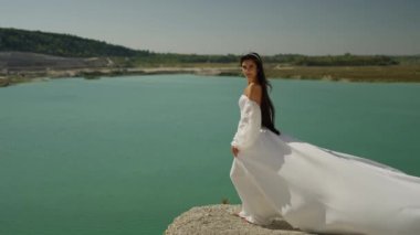 Beyaz gelinlik ve duvak giymiş genç gelinin muhteşem manzarası denizin veya okyanusun arka planında mavi suyla duran kayanın üzerinde duruyor..