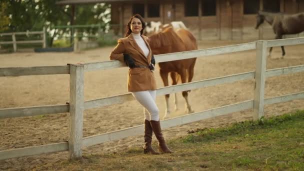 ファッショナブルでエレガントな服を着た若い女性は 牧場で放牧する馬とパドックの近くに立っています — ストック動画