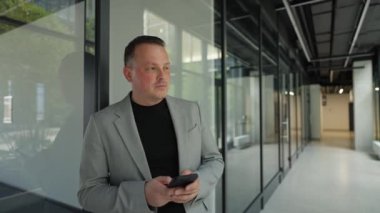 Modern bir iş binasında dururken akıllı telefonuyla kitap okuyan beyaz bir iş adamı.