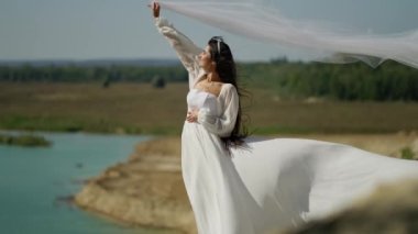 Beyaz elbiseli romantik gelin terasta poz veriyor. Deniz, dağlar ve rüzgâr peçesini açıyor.