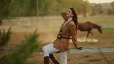 Genç ve sakin bir kadın çiftlikte dinlenirken ve ata binerken manzaranın ve atların tadını çıkarıyor.