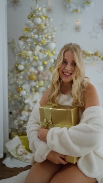 新しい年の贈り物を受け取る魅力的なかわいい若い女性の肖像画 クリスマスツリーの背景に対して家の家でクリスマスイブを祝うギフトボックスを保持する幸せな笑顔の女性 — ストック動画