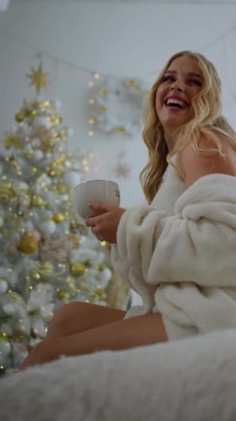 自宅で装飾されたクリスマスツリーの近くでホットドリンクでマグカップを保持しながら自信を持って微笑む若い金髪の女性 — ストック動画