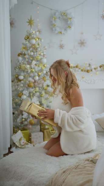 装飾されたクリスマスツリーの近くのリビングルームに座っている間クラフト紙にプレゼントを保持する魅力的な若い金髪の女性の肖像画 冬のお祝いの気分 — ストック動画