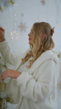 Çekici genç bir kadın parlak bir oturma odasındaki bir dala yılbaşı oyuncağı koydu. Çekici sarışın kadın gülümsüyor ve Noel ağacını süslüyor..