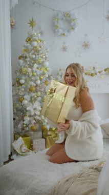 Çekici, kıvırcık saçlı beyaz bir kadın içinde mutlu bir sürpriz olan şirin bir hediye kutusunu sallıyor. İçinde ne olduğunu tahmin etmeye çalışıyor. Köknar ağacının arka planında kameraya gülümsüyor.