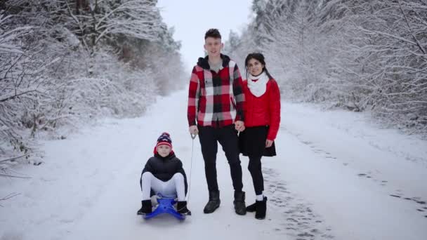 冬の雪の公園でソリと一緒に歩いている小さな息子と幸せな夫婦 若い家族 小さな息子を持つ夫と妻 週末に一緒にレジャー時間を過ごす — ストック動画