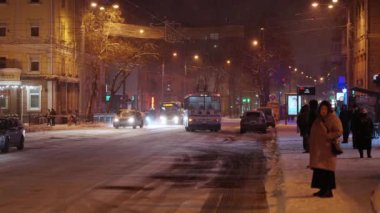Caddelerde yoğun kar var, kar fırtınası var, insanlar otobüs durağında otobüs bekliyor, yollarda trafik sıkışıklığı var.
