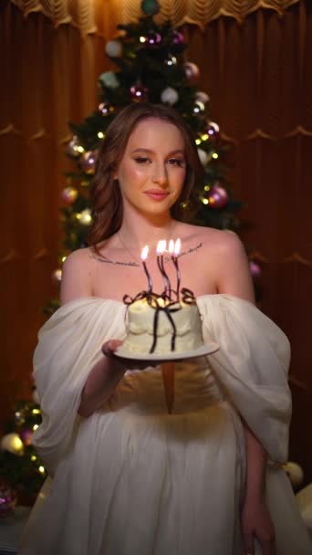 快乐微笑的年轻女子拿着生日蛋糕 在一棵装饰着灿烂花环的圣诞树前吹灭了一支蜡烛 — 图库视频影像