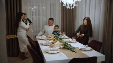 Genç bir çift ve sevimli küçük çocuklar yemek masasında birlikte oturup ailemle kutlama yaparken gülümseyip kameraya bakıyorlar.